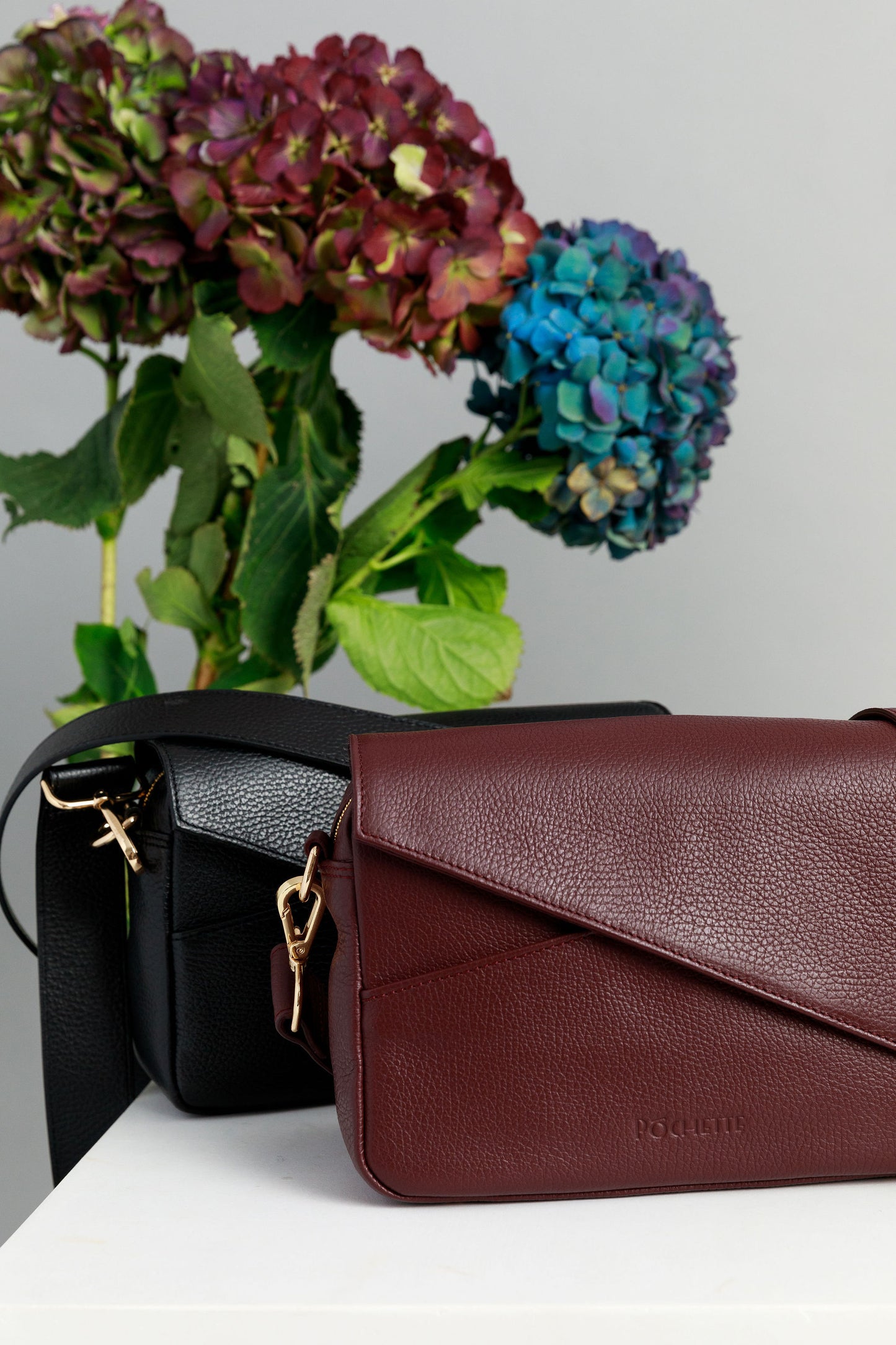Black and burgundy crossbody bags on a white surface with wide leather straps attached. A bunch of three hydrangeas in burgundy, blue and purple are in the background.