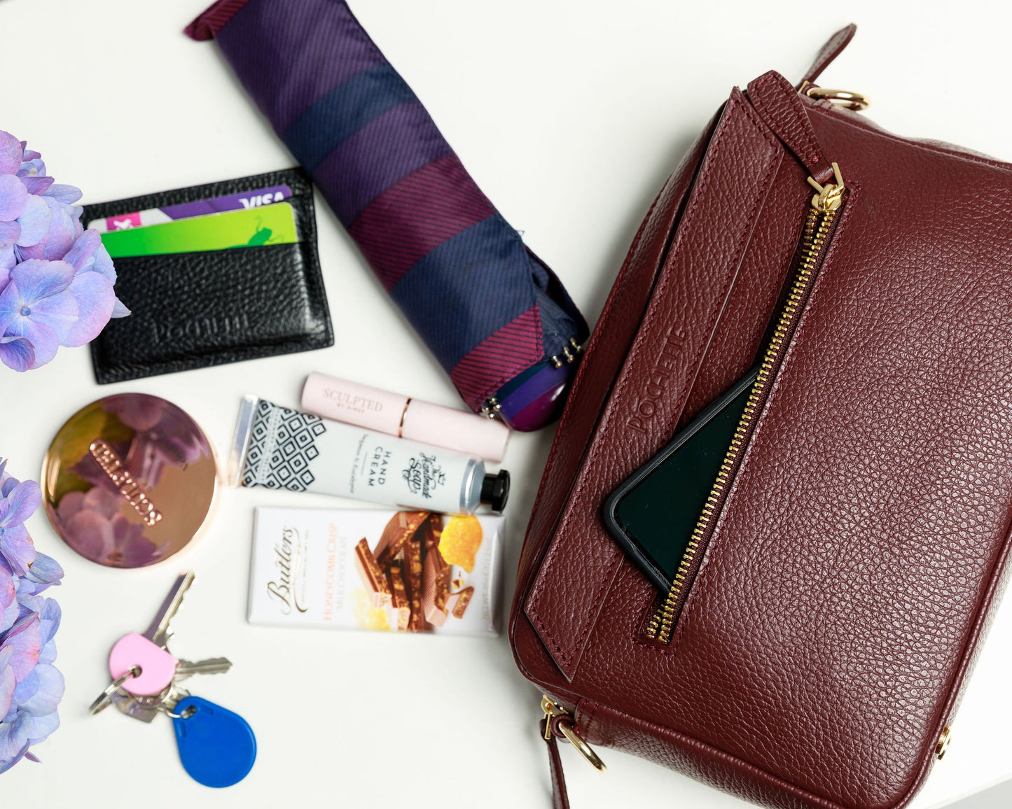 A burgundy crossbody bag on a white surface with blurred hydrangeas in the foreground. The contents of the bag are shown to be spilling out of the handbag, which can fit a mobile phone, handcream from the Homemade Soap Company, lipstick and face powder from Sculpted by Aimee, a black Póchette wallet, keys, an umbrella and some Butler's chocolate for emergencies.