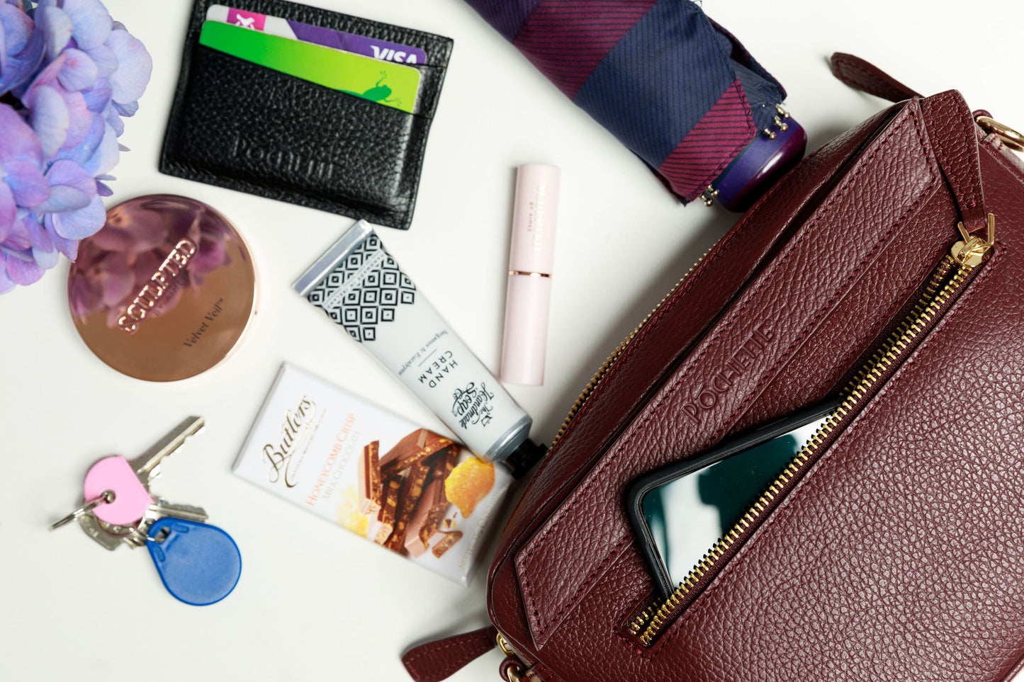 A burgundy crossbody bag on a white surface with blurred hydrangeas in the foreground. The contents of the bag are shown to be spilling out of the handbag, which can fit a mobile phone, handcream from the Homemade Soap Company, lipstick and face powder from Sculpted by Aimee, a black Póchette wallet, keys, an umbrella and some Butler's chocolate for emergencies.