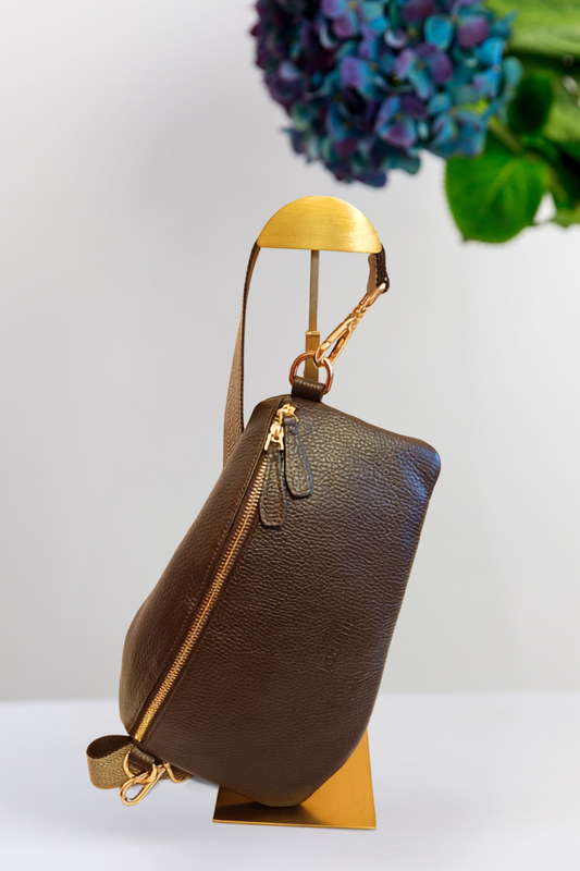 A dark brown pebbled leather shoulder bag with gold-tone hardware and a woven strap, displayed on a gold-colored stand against a white background with a blurred purple flower in the upper right.
