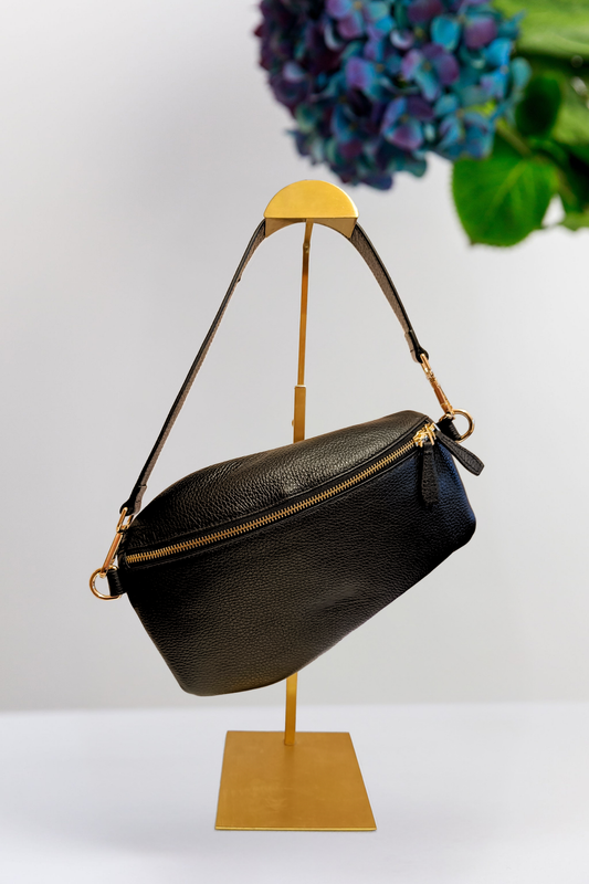 A black pebbled leather shoulder bag with gold-tone hardware, displayed on a gold metal stand against a white background with a blurred floral arrangement in the upper right.