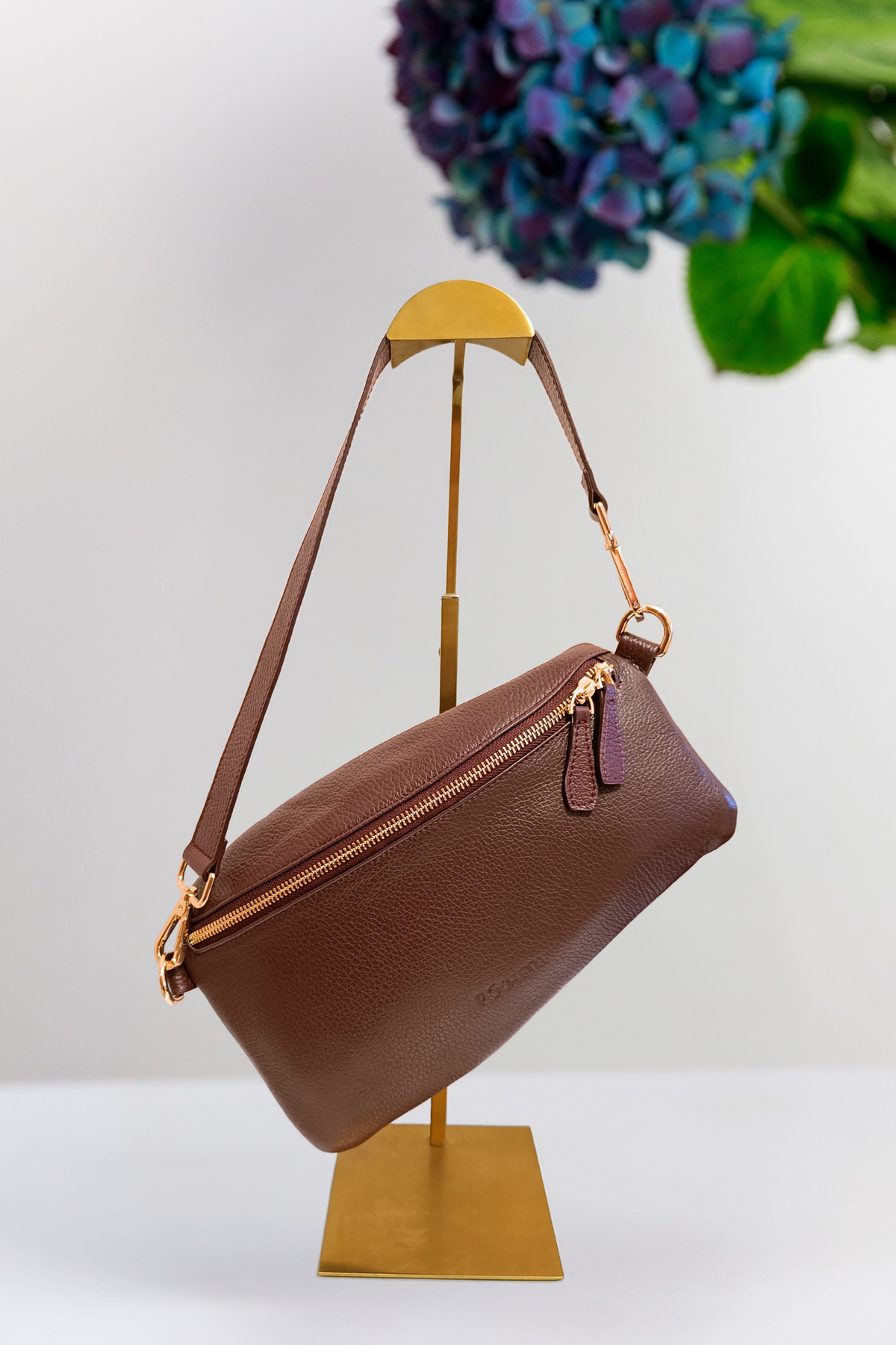 A burgundy pebbled leather shoulder bag with gold-tone hardware, displayed on a gold metal stand against a white background with a blurred floral arrangement in the upper right.