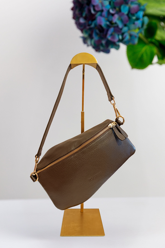 A chocolate brown pebbled leather shoulder bag with gold-tone hardware, displayed on a gold metal stand against a white background with a blurred floral arrangement in the upper right.