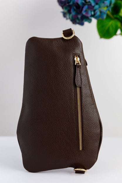 A brown leather swing crossbody bag with a zipper and a metallic clasp, displayed against a white background with a floral pattern in the upper right corner.