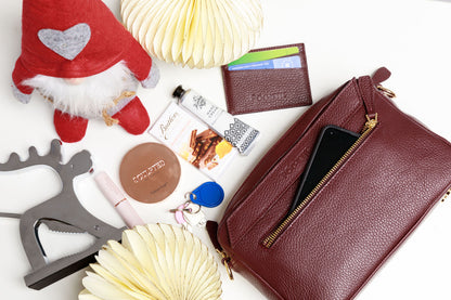 A burgundy crossbody bag on a white surface with christmas decorations. The contents of the bag are shown to be spilling out of the handbag, which can fit a mobile phone, handcream from the Homemade Soap Company, lipstick and face powder from Sculpted by Aimee, a black Póchette wallet, keys, an umbrella and some Butler's chocolate for emergencies. 