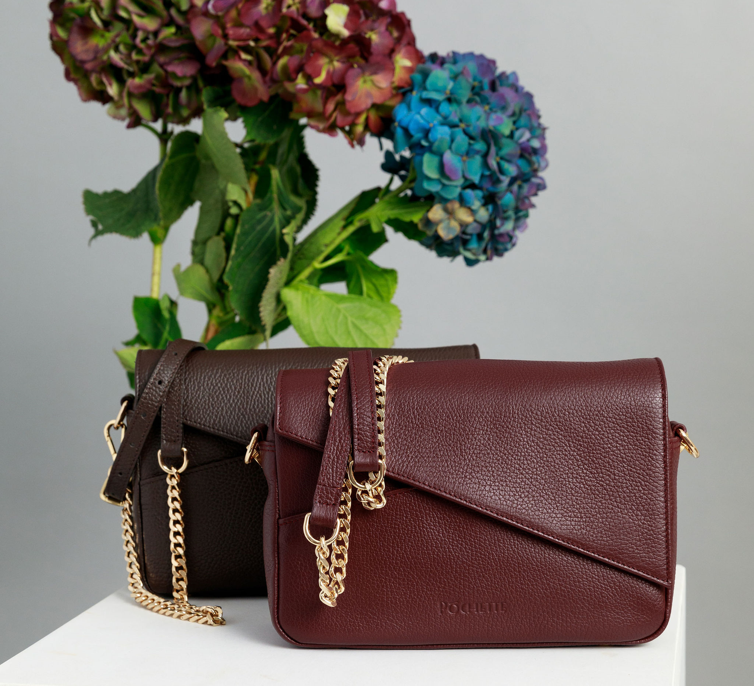 Chocolate and burgundy crossbody bags on a white surface with leather and chain straps attached. A bunch of three hydrangeas in burgundy, blue and purple are in the background.