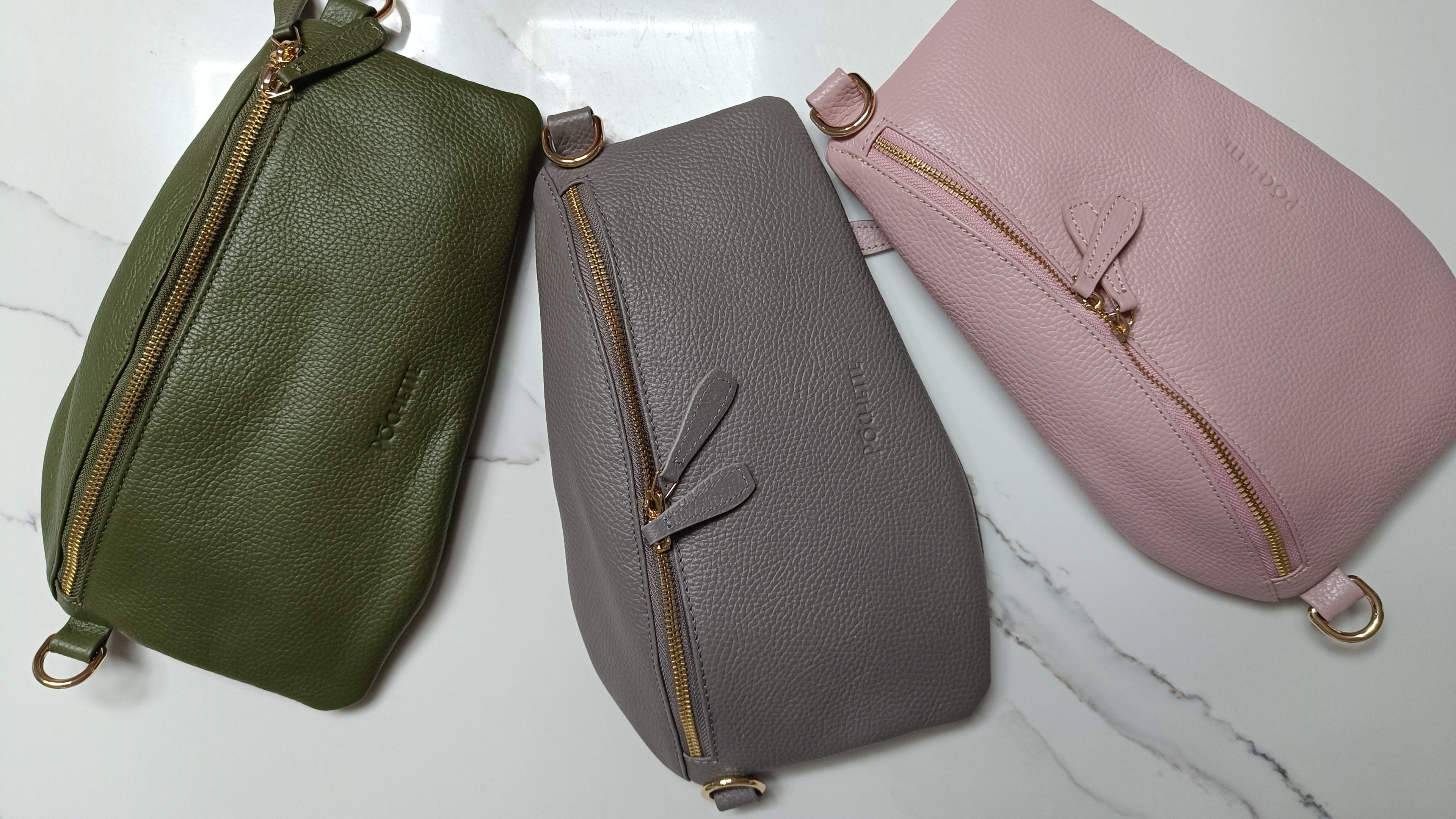 Three Póca crossbody bags - one in olive green, one in grey and one in powder pink on a white and grey marble counter.