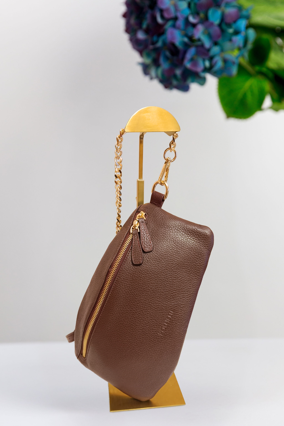 A burgundy pebbled leather bag with gold-toned hardware and a chain strap, displayed on a gold stand against a white background with a blurred purple flower in the upper right.