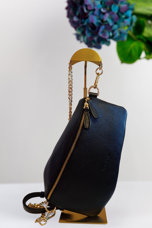 A black pebbled leather shoulder bag with gold-tone hardware and a chain strap, displayed on a gold stand against a white background with a blurred floral arrangement in the upper right.