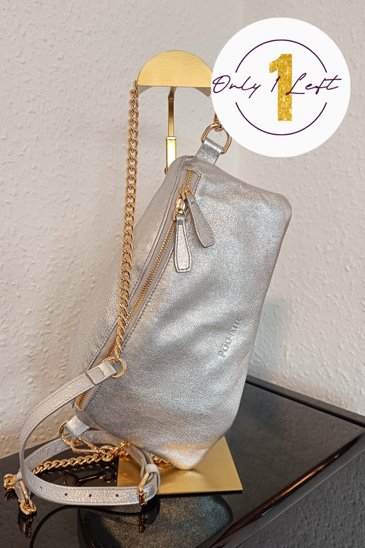 A metallic silver shoulder bag with gold chain straps, displayed on a gold stand against a textured off-white wall. A white circular sticker overlay in the top right corner features the text 'Only 1 Left' in purple and gold.