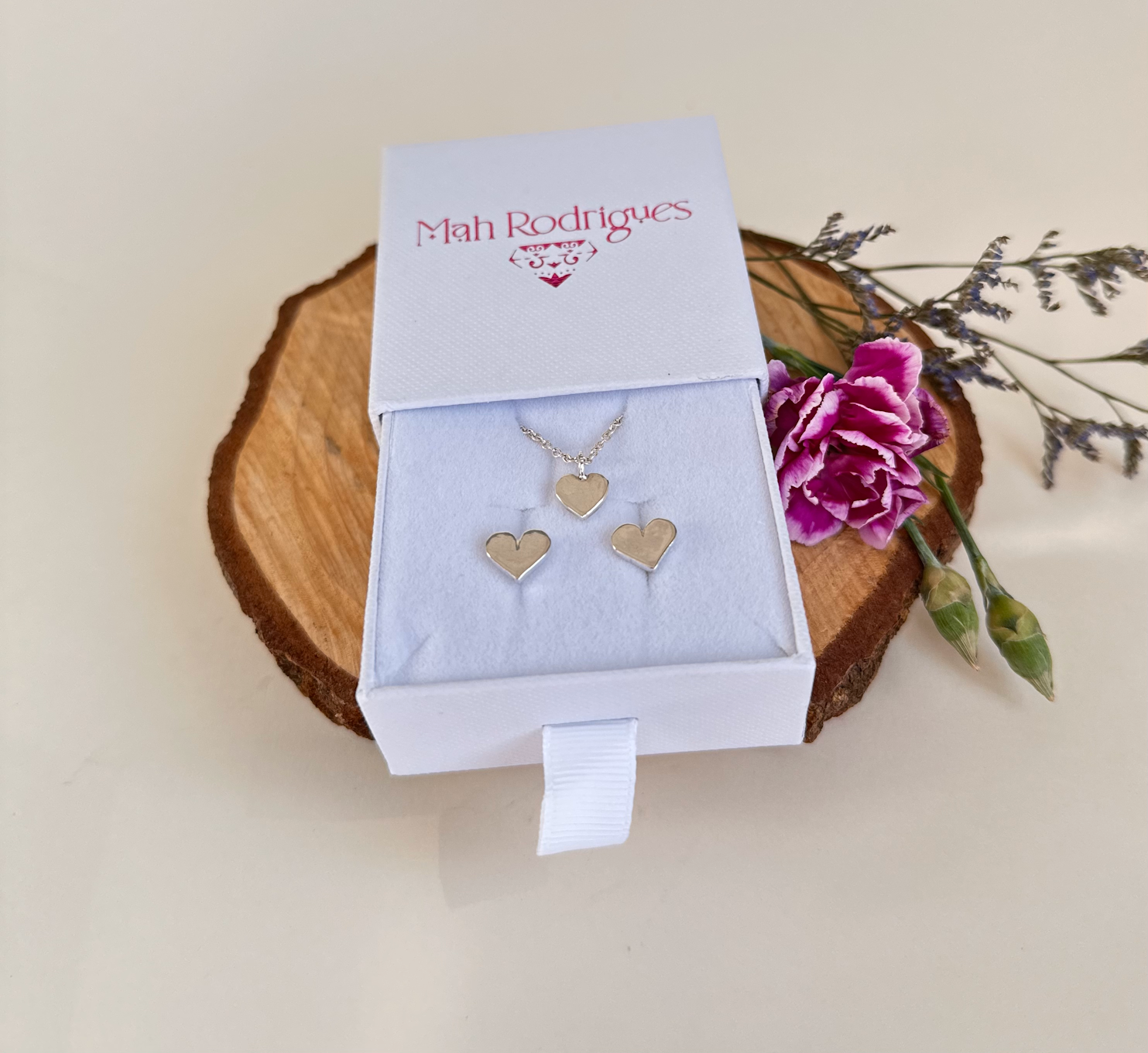 A photos of a heart pendant on a chain with matching heart earrings in silver in a white box on a piece of tree  with some flowers