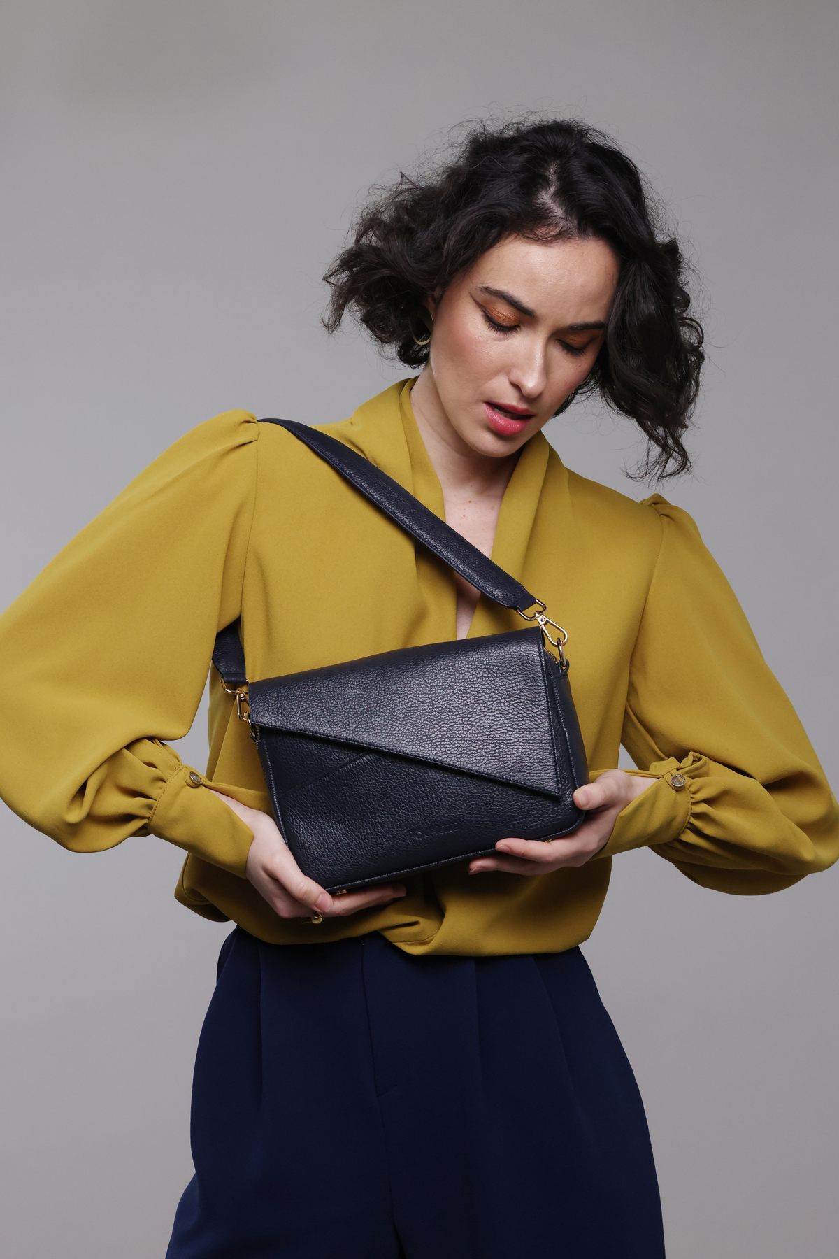 A dark haired woman wearing a mustard yellow blouse, navy pants and a navy blue leather handbag