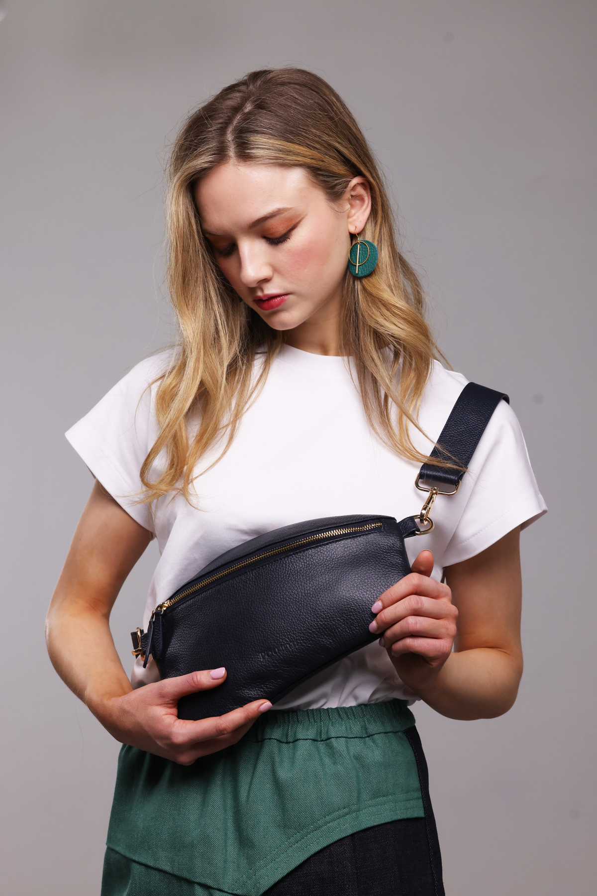 A blonde haired woman wearing a white t-shirt, a teal and denim skirt and a navy blue leather crossbody bag.