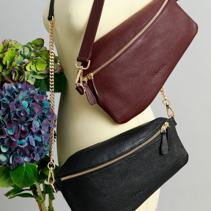 A burgundy Póca swing bag with a short leather shoulder strap and a black Póca swing bag with a leather and chain strap on a sewing mannequin. A bunch of three hydrangeas in burgundy, blue and purple are beside it.