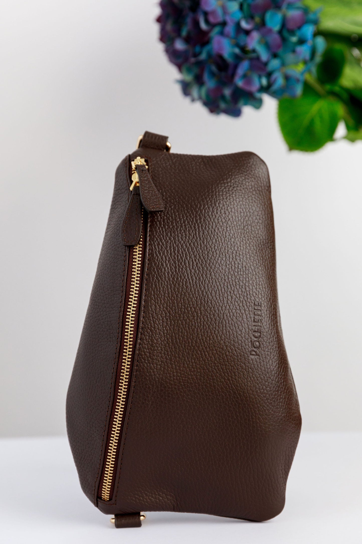 A chocolate brown swing crossbody bag on a white surface with blurred hydrangeas in the background.