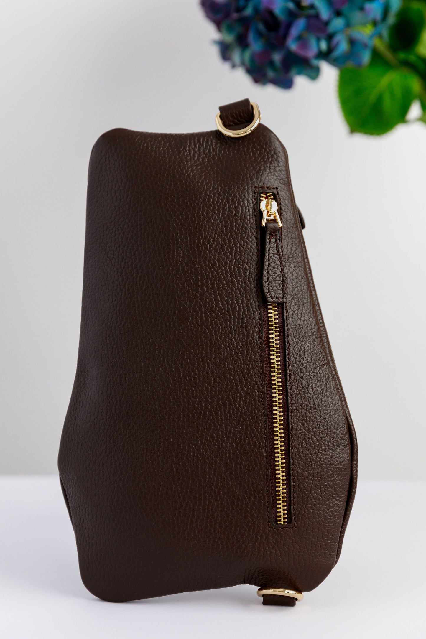 The back of a chocolate brown swing crossbody bag, called the Póca, on a white surface with blurred hydrangeas in the background.