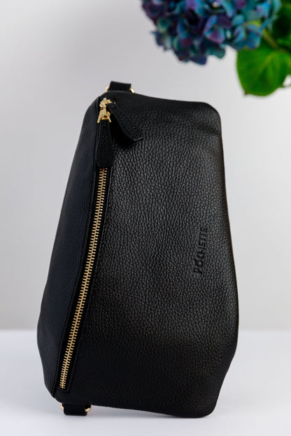 A black swing crossbody bag, called the Póca, on a white surface with blurred hydrangeas in the background.