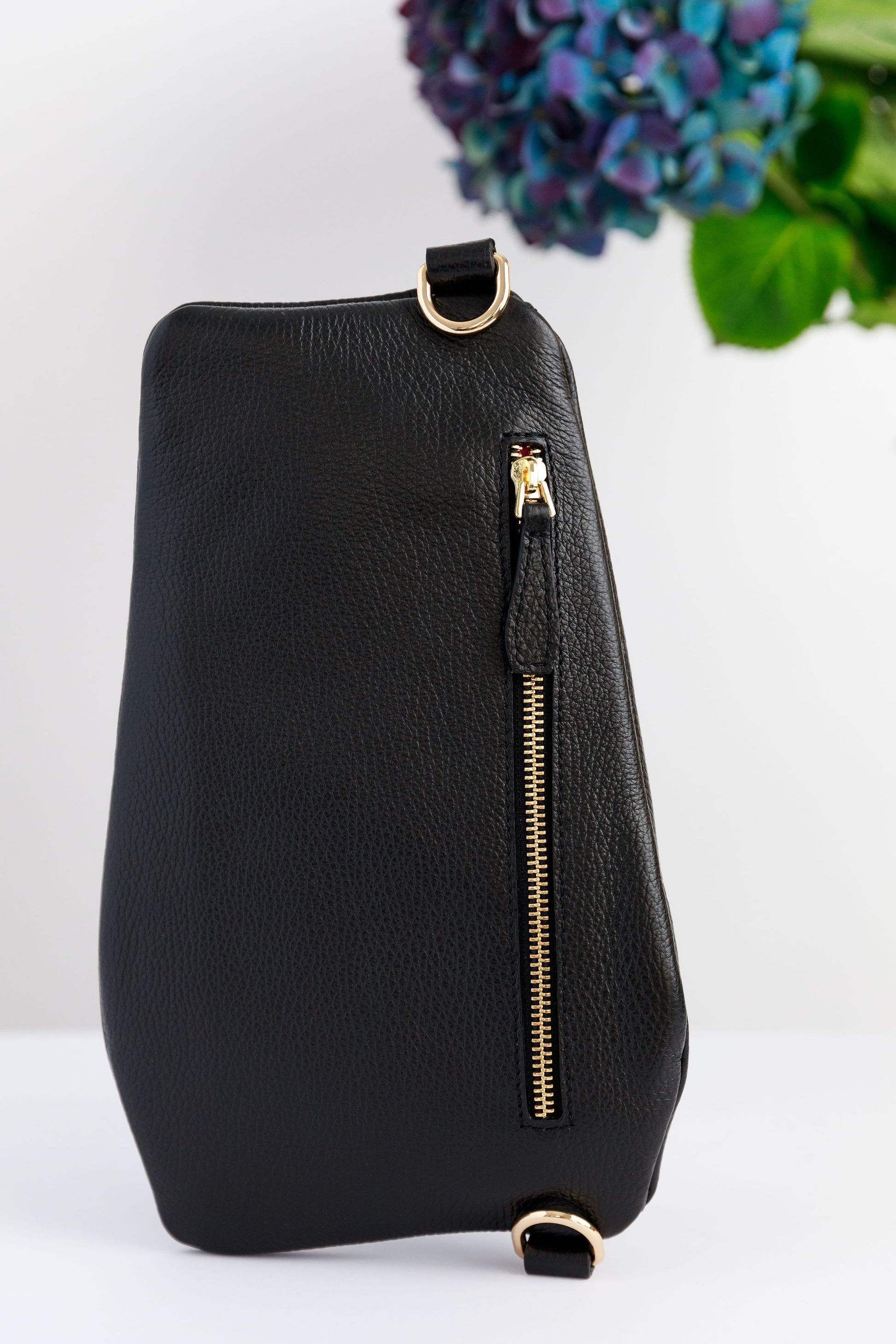 The back of a black swing crossbody bag, called the Póca, on a white surface with blurred hydrangeas in the background.