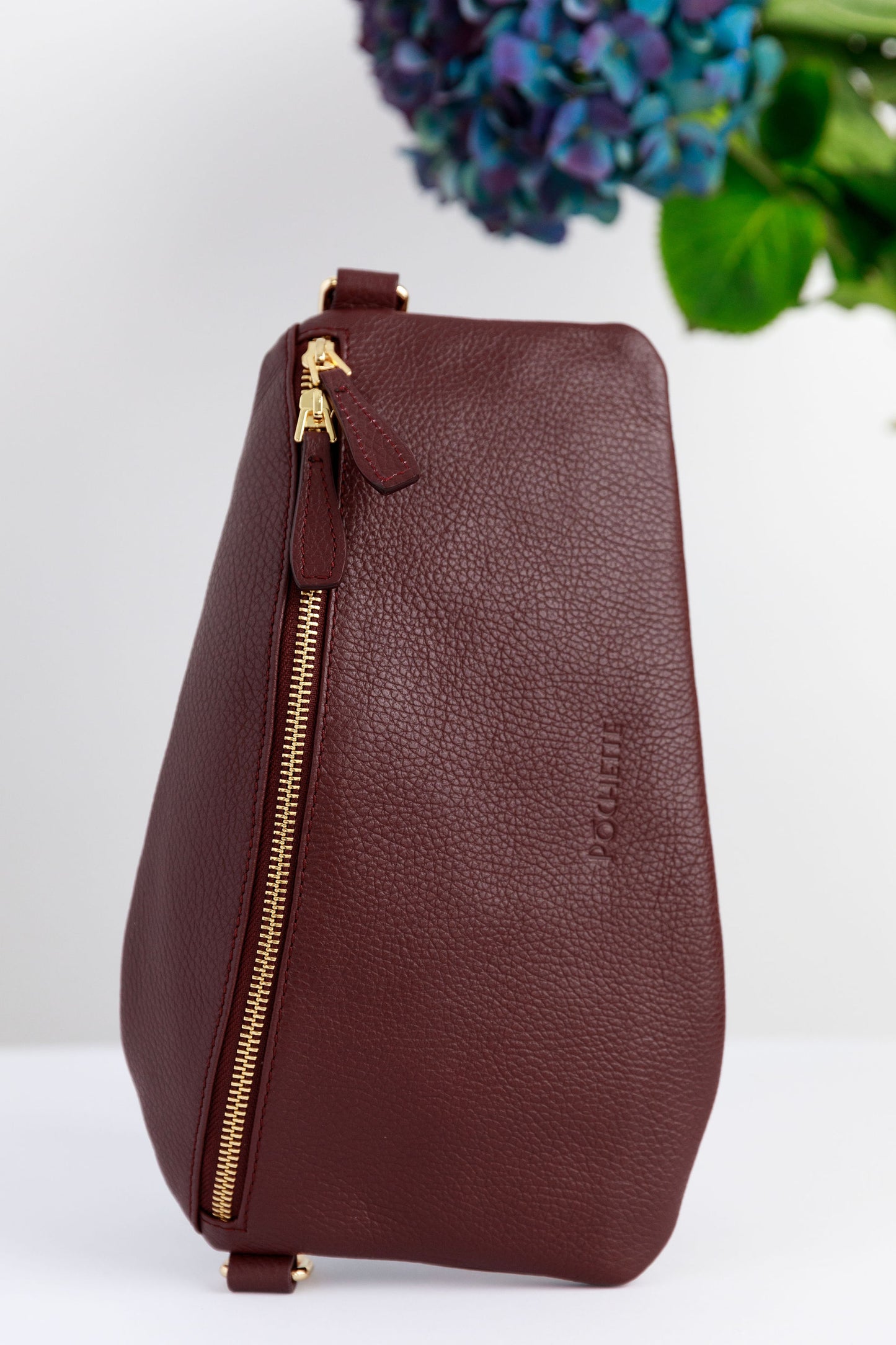 A burgundy swing crossbody bag on a white surface with blurred hydrangeas in the background.