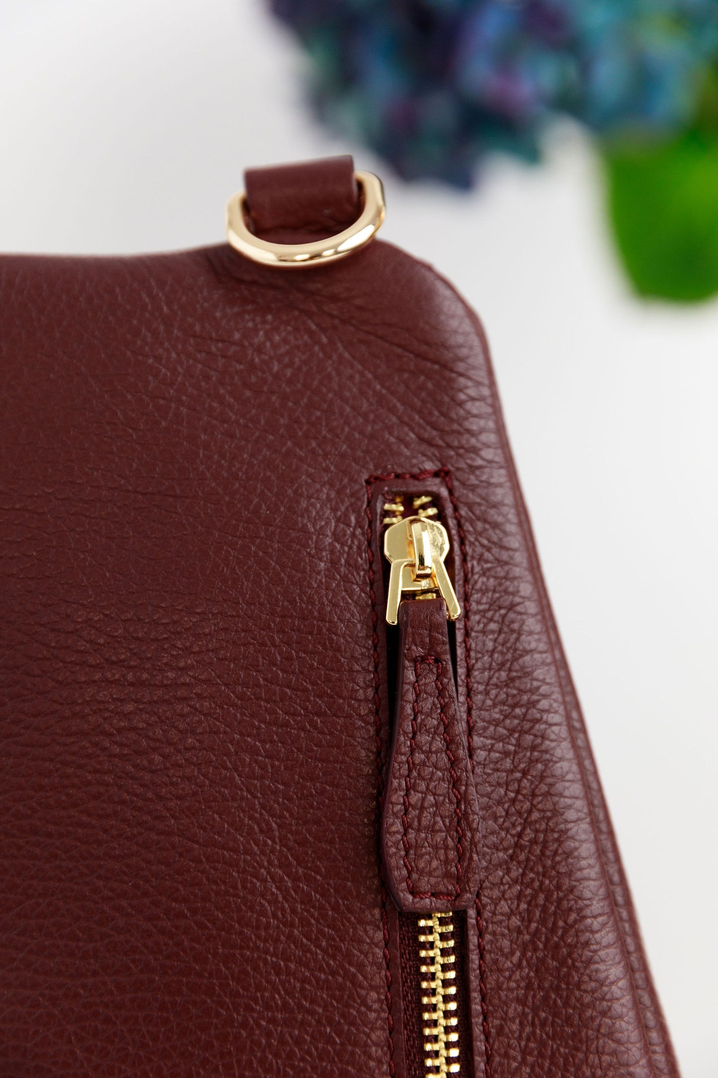 A close up of the burgundy swing crossbody bag, called the Póca, with blurred hydrangeas in the background. The zipped pocket at the back and the Póchette shaped zipper pull are displayed.