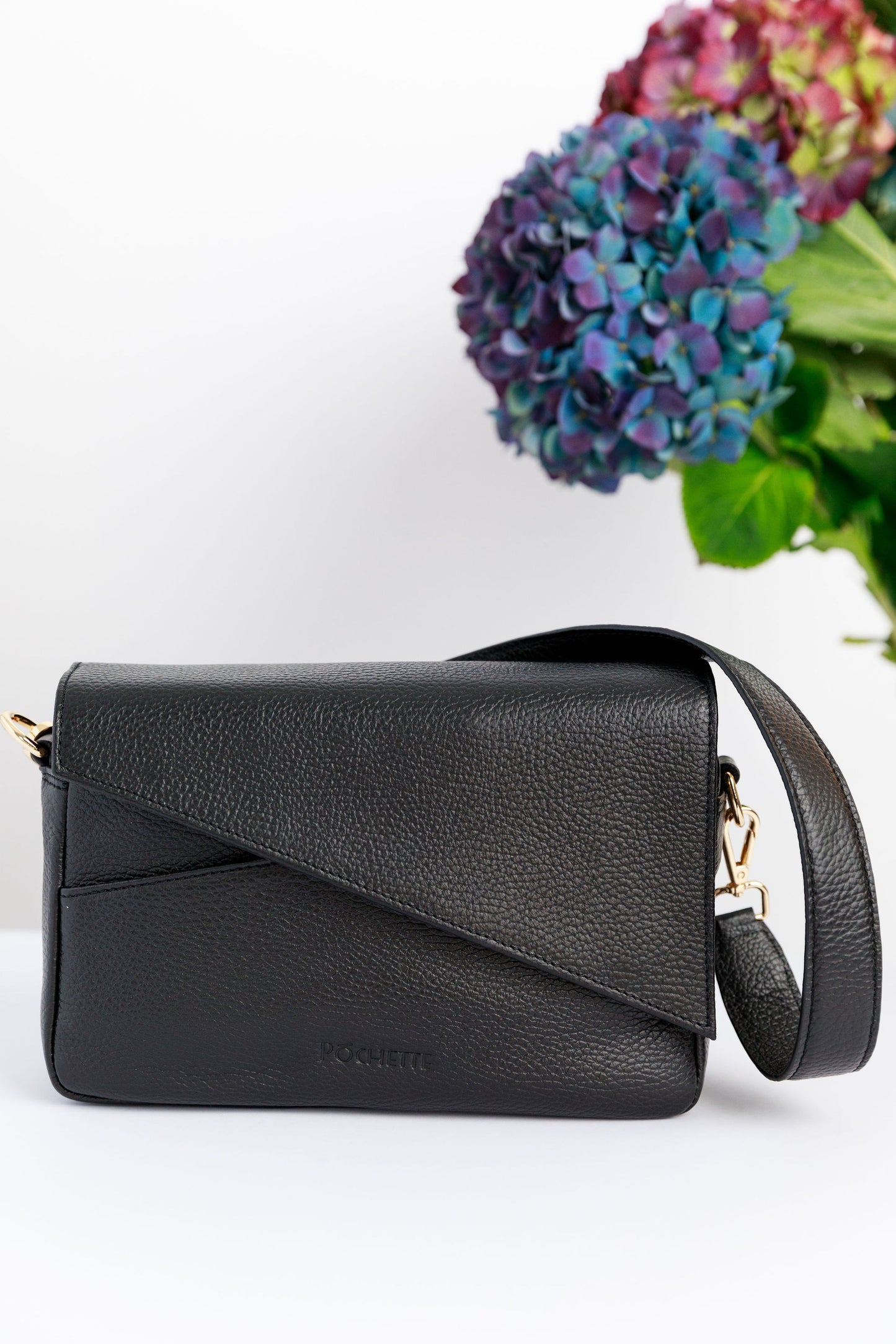 A black crossbody bag on a white surface with blurred hydrangeas in the background.