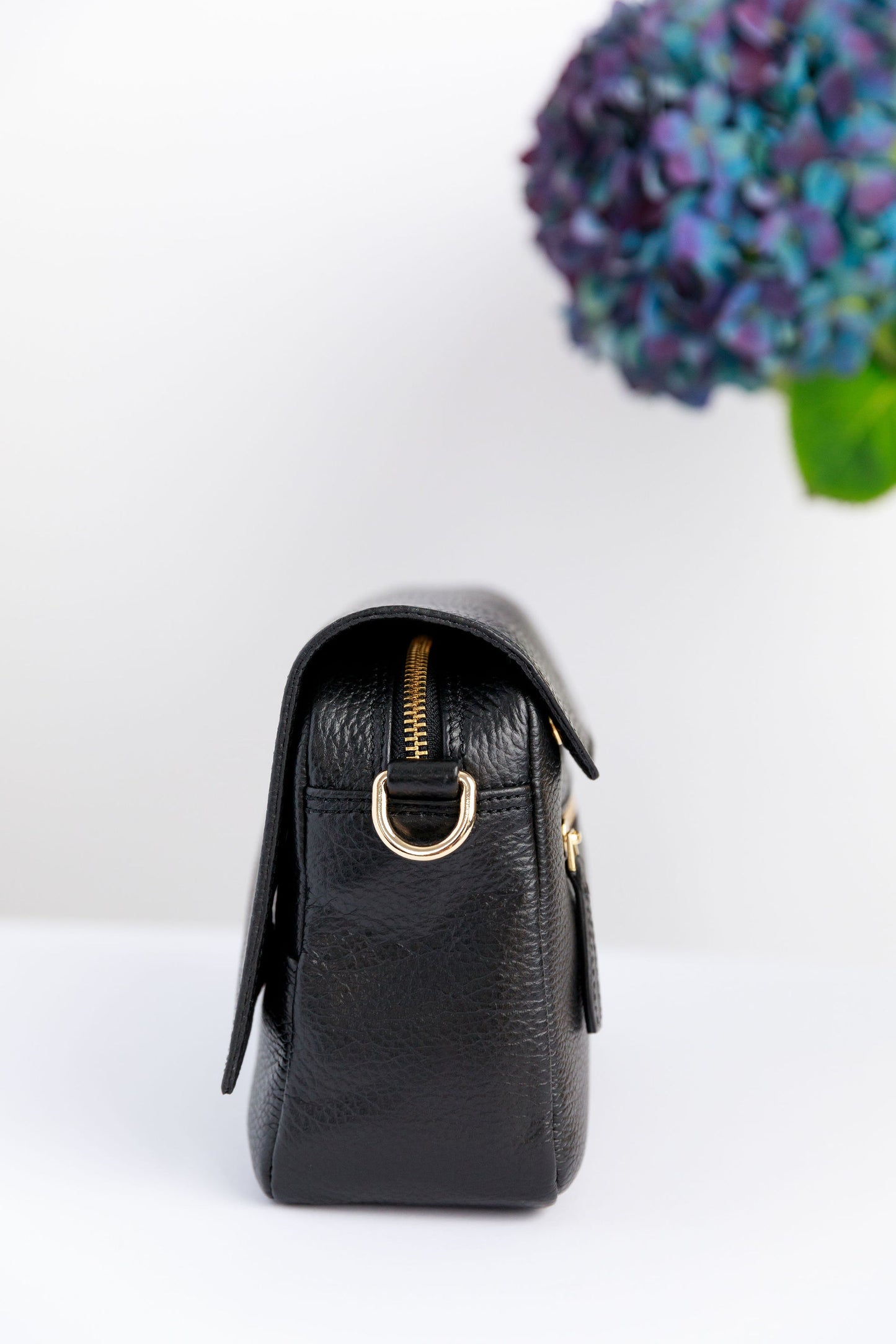 The side of a black crossbody bag on a white surface with blurred hydrangeas in the background.