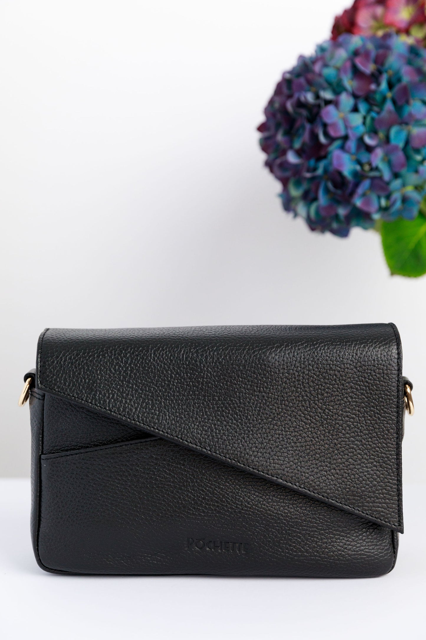 A black crossbody bag on a white surface with blurred hydrangeas in the background.