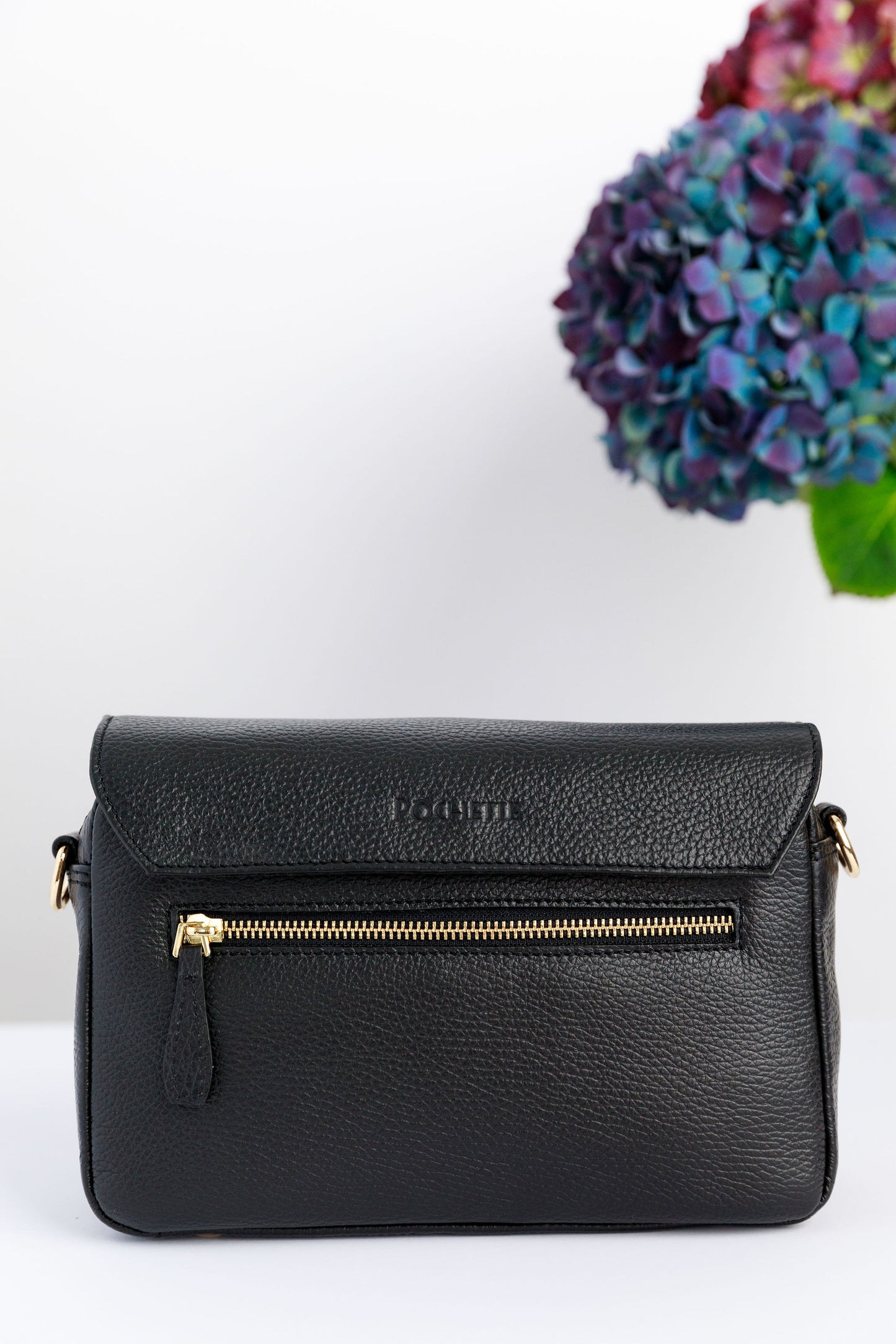 The back of a black crossbody bag on a white surface with blurred hydrangeas in the background.