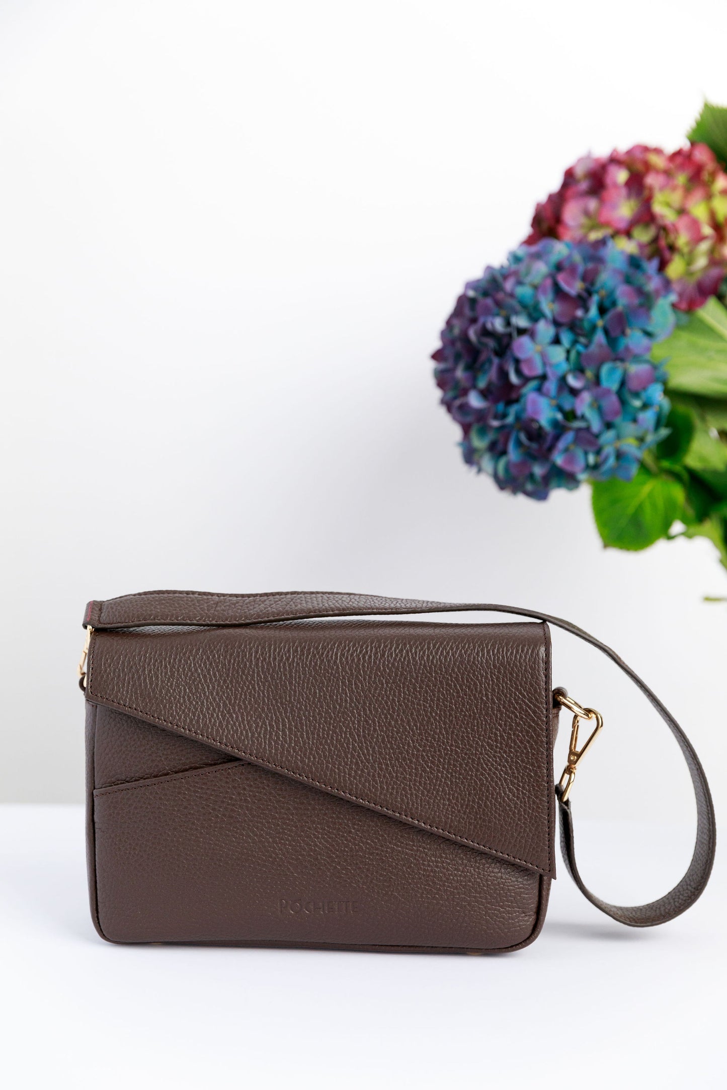 A chocolate brown crossbody bag on a white surface with blurred hydrangeas in the background.