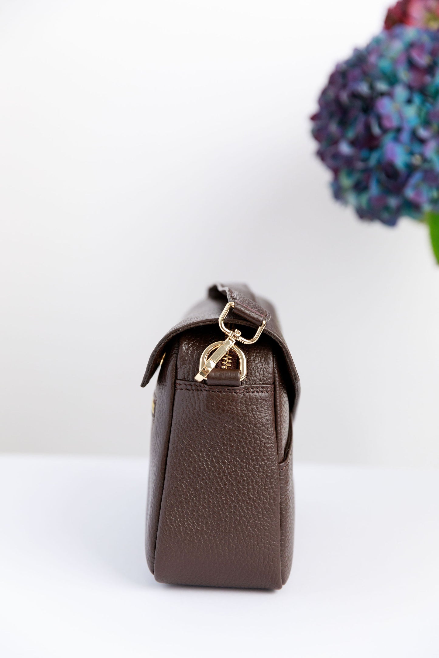 The side of a chocolate brown crossbody bag on a white surface with blurred hydrangeas in the background.