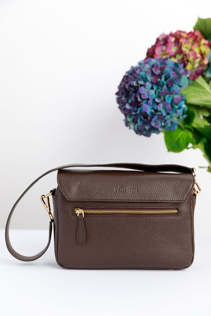 A back of a chocolate brown crossbody bag on a white surface with blurred hydrangeas in the background.