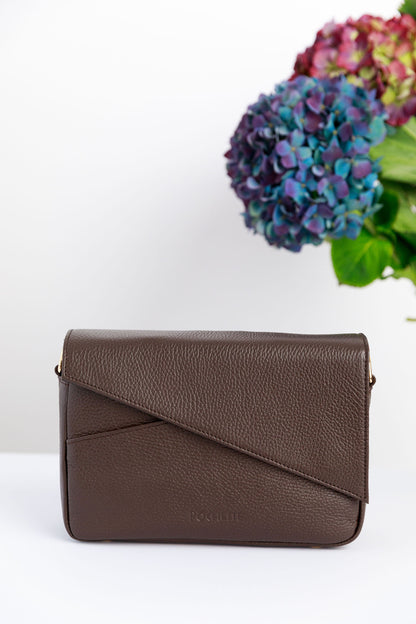 A chocolate brown crossbody bag on a white surface with blurred hydrangeas in the background.