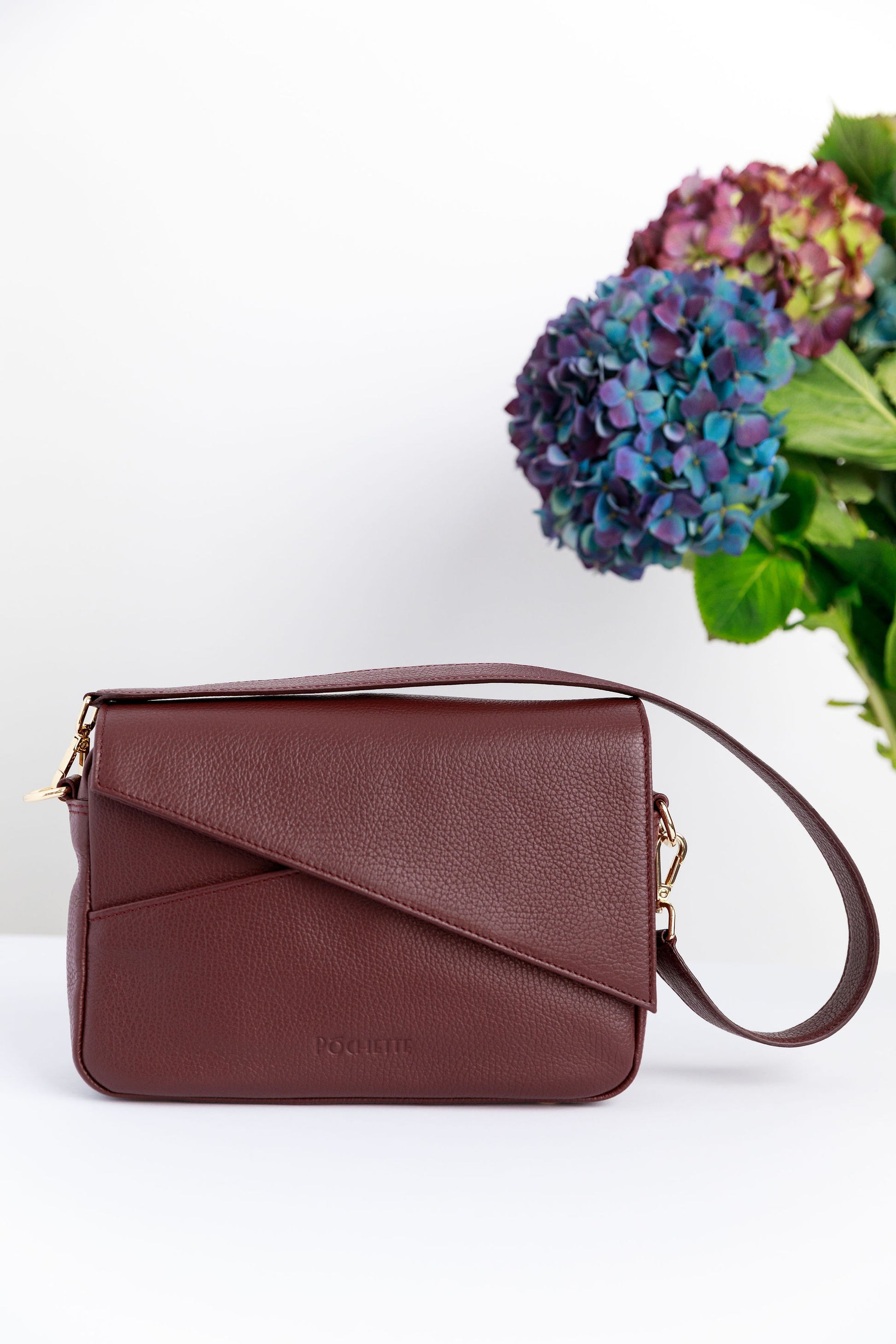 A burgundy crossbody bag on a white surface with blurred hydrangeas in the background.