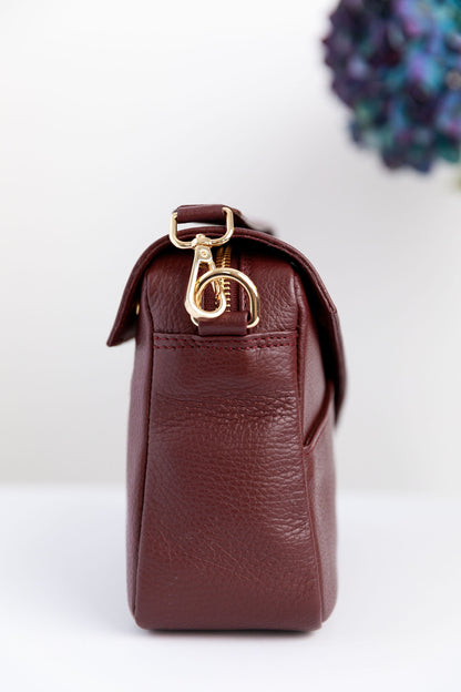 The side of a burgundy crossbody bag on a white surface with blurred hydrangeas in the background.