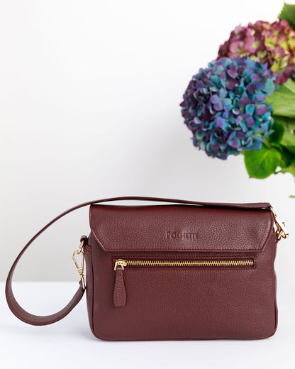 The back of a burgundy crossbody bag on a white surface with blurred hydrangeas in the background.