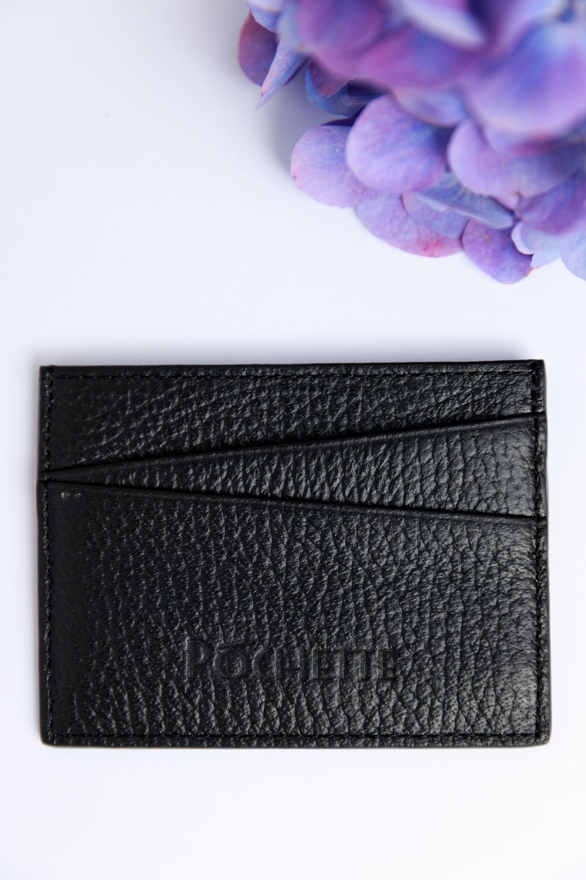 A black card wallet on a white surface with a blurred hydrangea in the foreground.