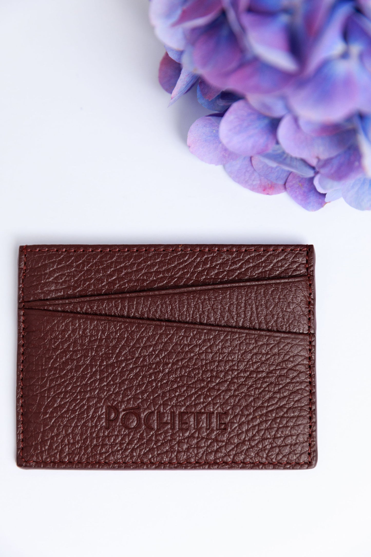 A burgundy card wallet on a white surface with a blurred hydrangea in the foreground.