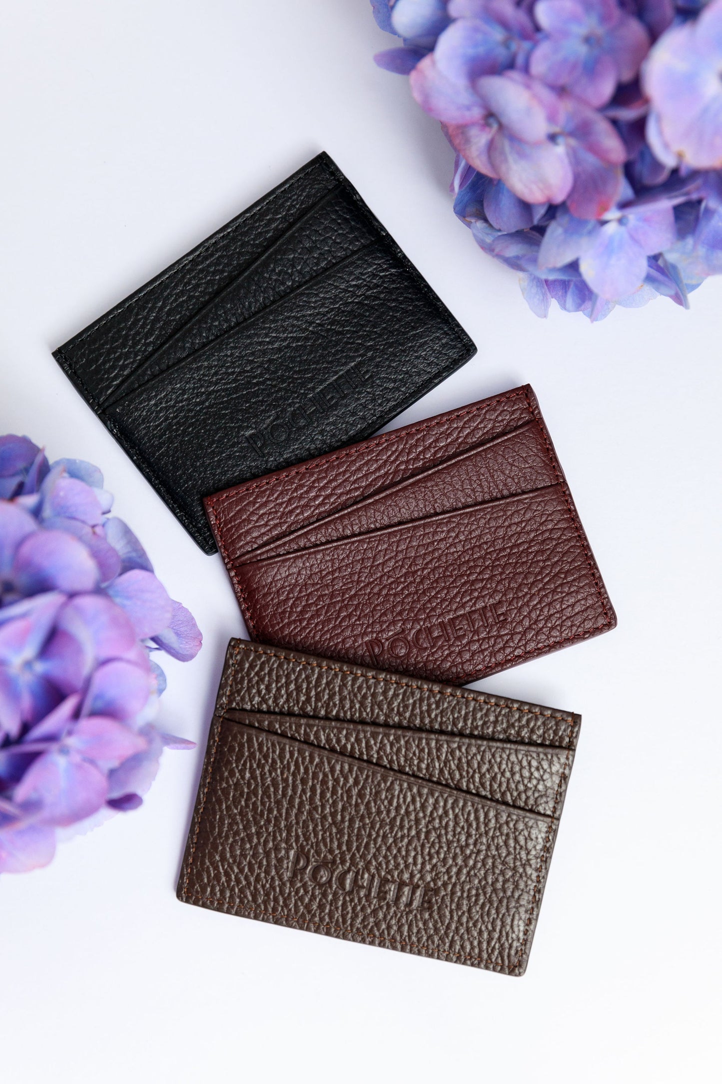 Black, burgundy and choclate brown card wallets on a white surface with a blurred hydrangea in the foreground.