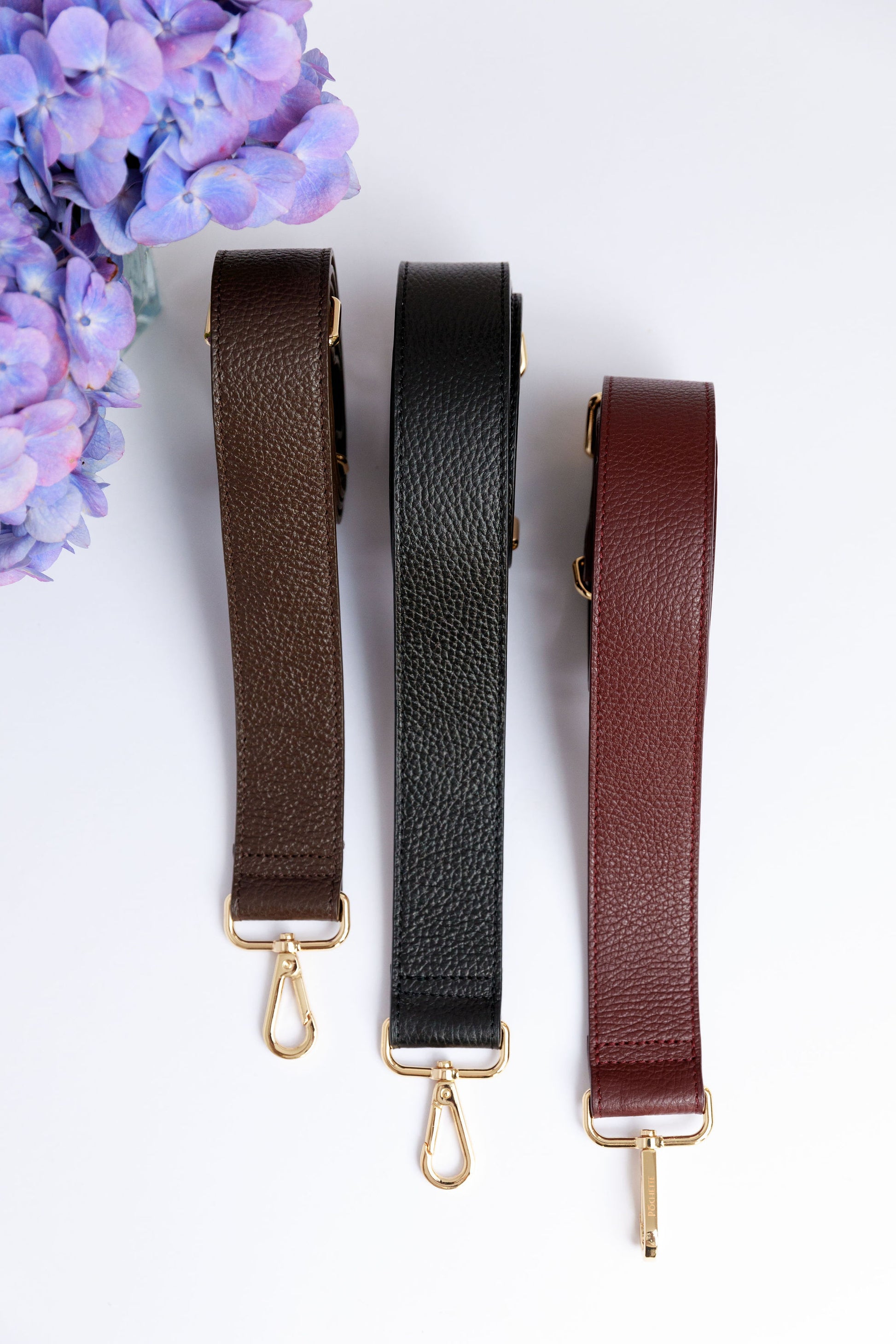 Three wide leather straps in burgundy, black and chocolate brown on a white surface with a blurred hydrangea in the foreground.