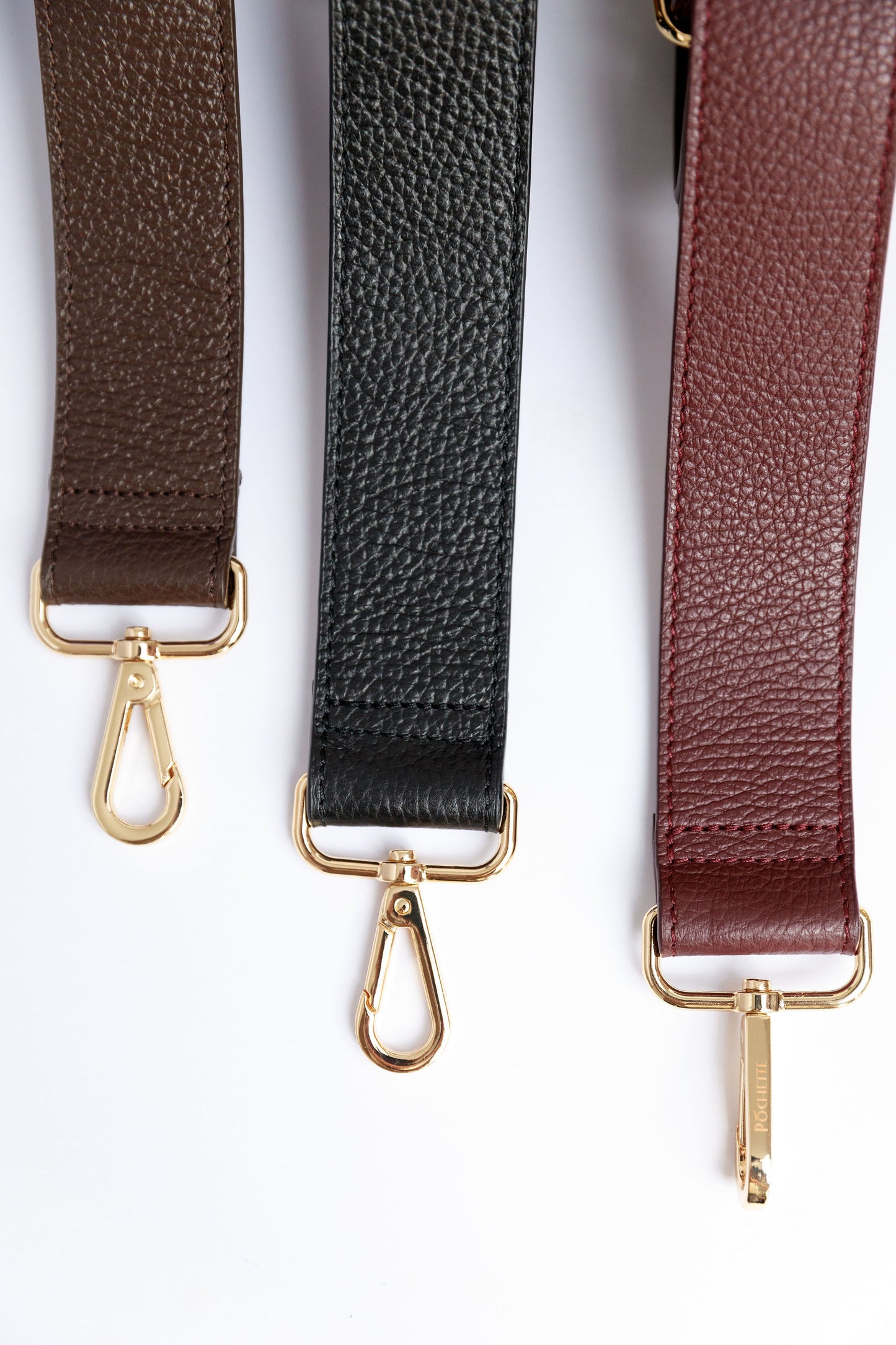 A close up of three wide leather straps in burgundy, black and chocolate brown on a white surface.