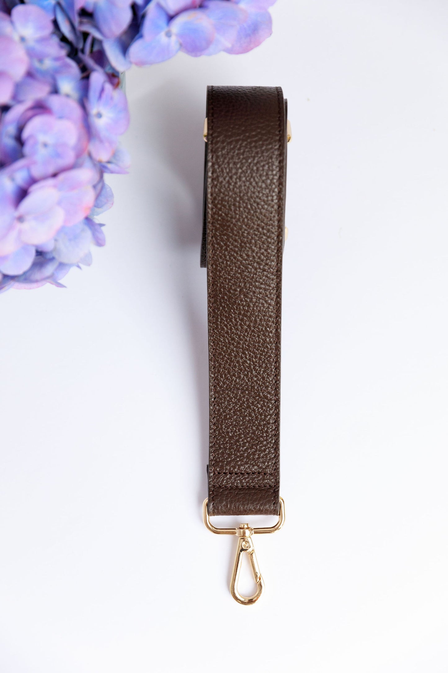 A chocolate brown wide leather strap on a white surface with a blurred hydrangea in the foreground.