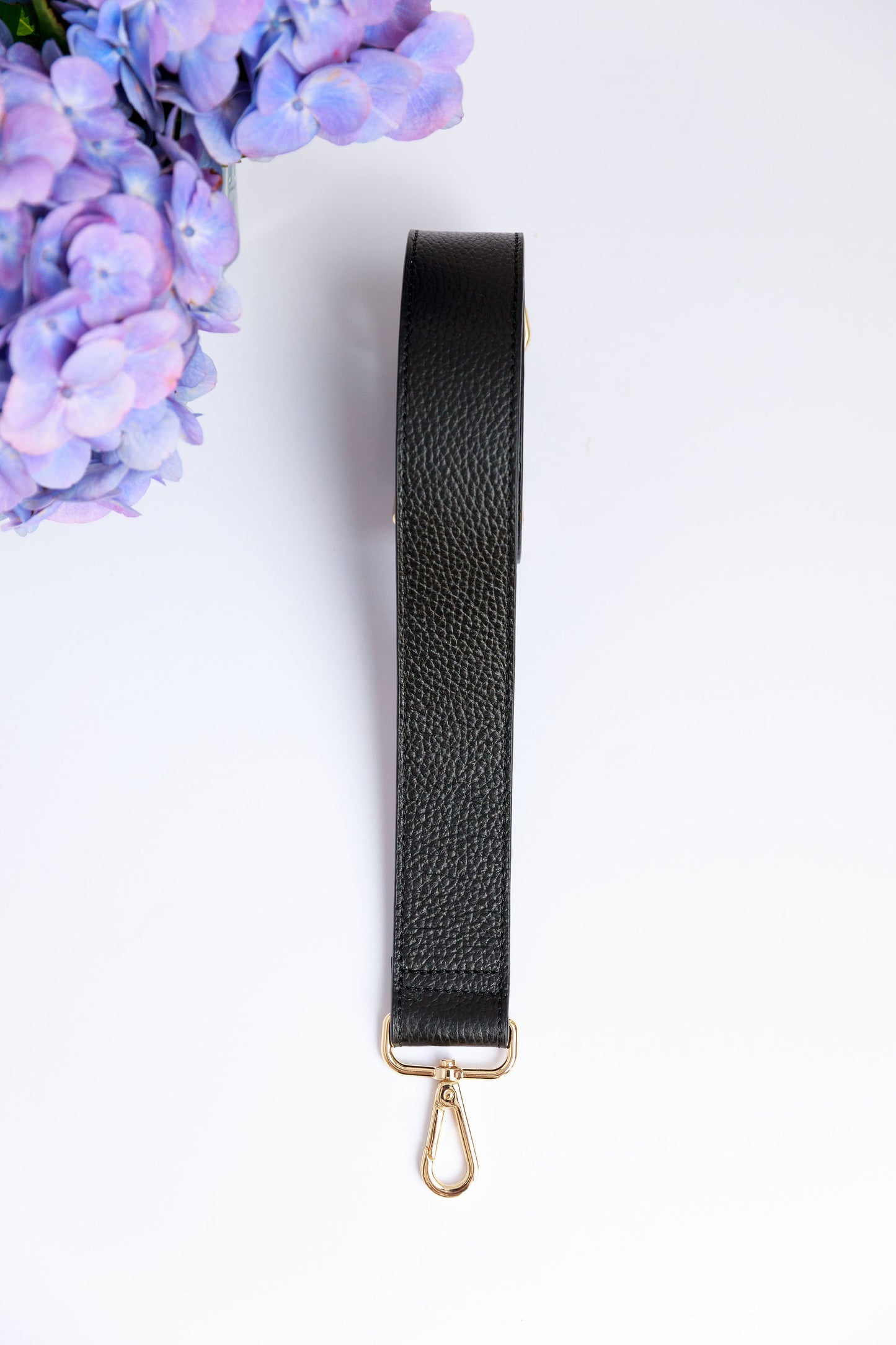 A black wide leather strap on a white surface with a blurred hydrangea in the foreground.