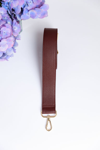 A burgundy wide leather strap on a white surface with a blurred hydrangea in the foreground.