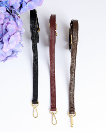 Three thin leather straps in burgundy, black and chocolate brown on a white surface with a blurred hydrangea in the foreground.