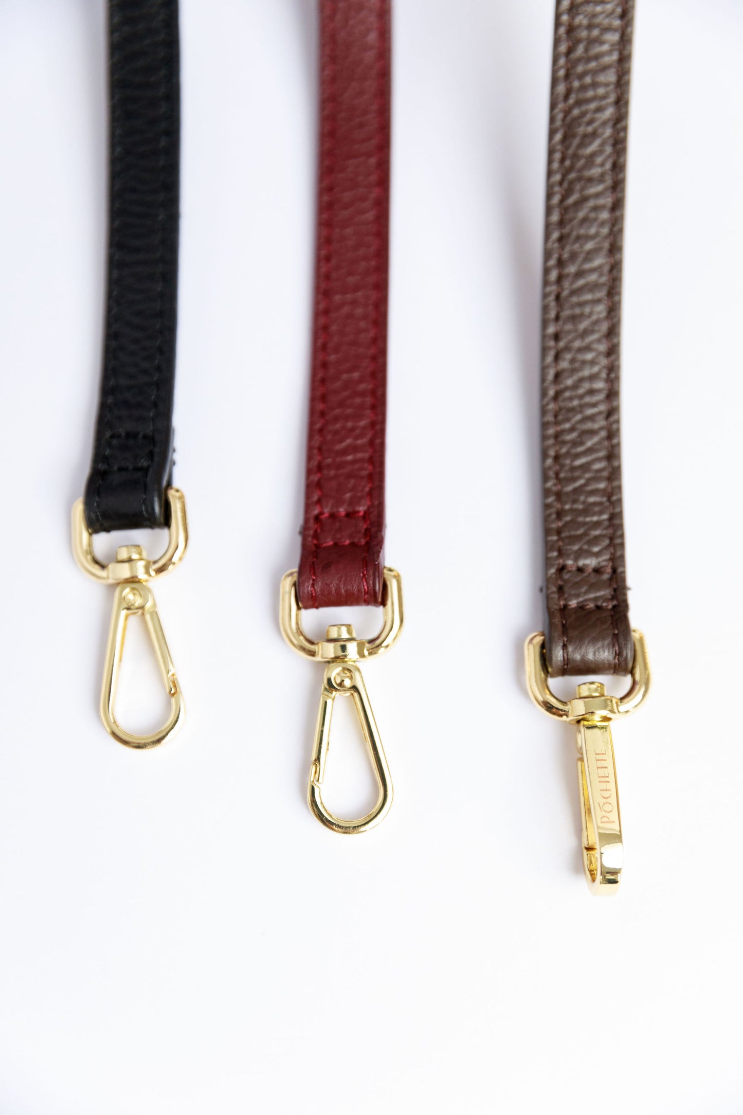 A close up of three thin leather straps in burgundy, black and chocolate brown on a white surface.