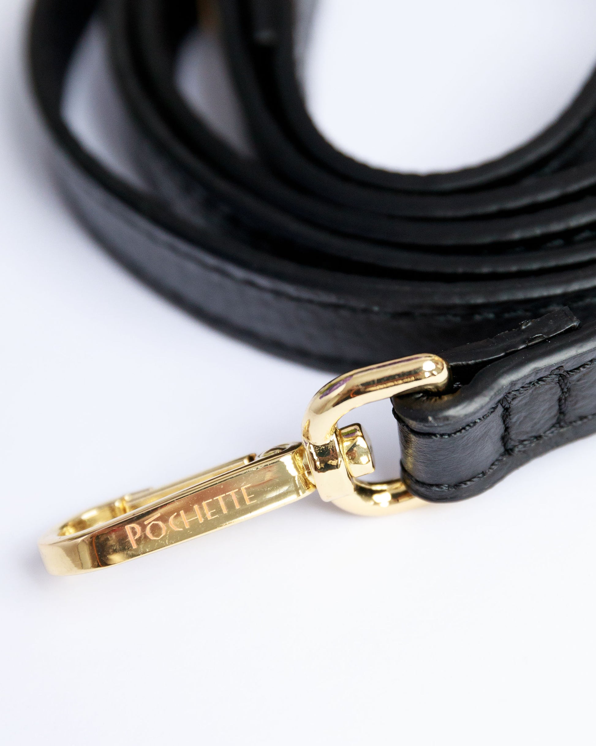 Close up of Póchette branding on the snap hook of a black, thin leather crossbody strap.