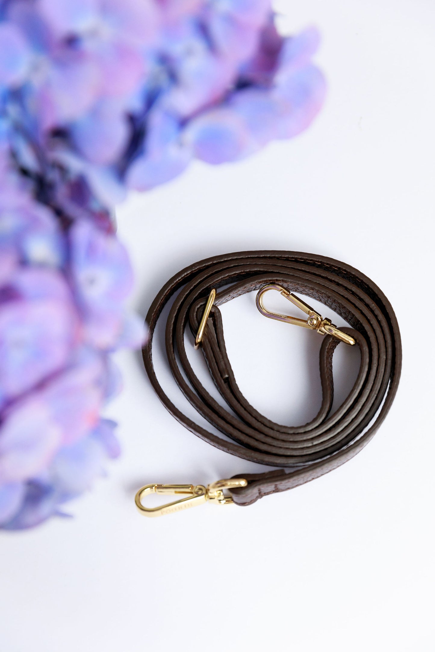 A chocolate brown thin leather strap on a white surface with a blurred hydrangea in the foreground.