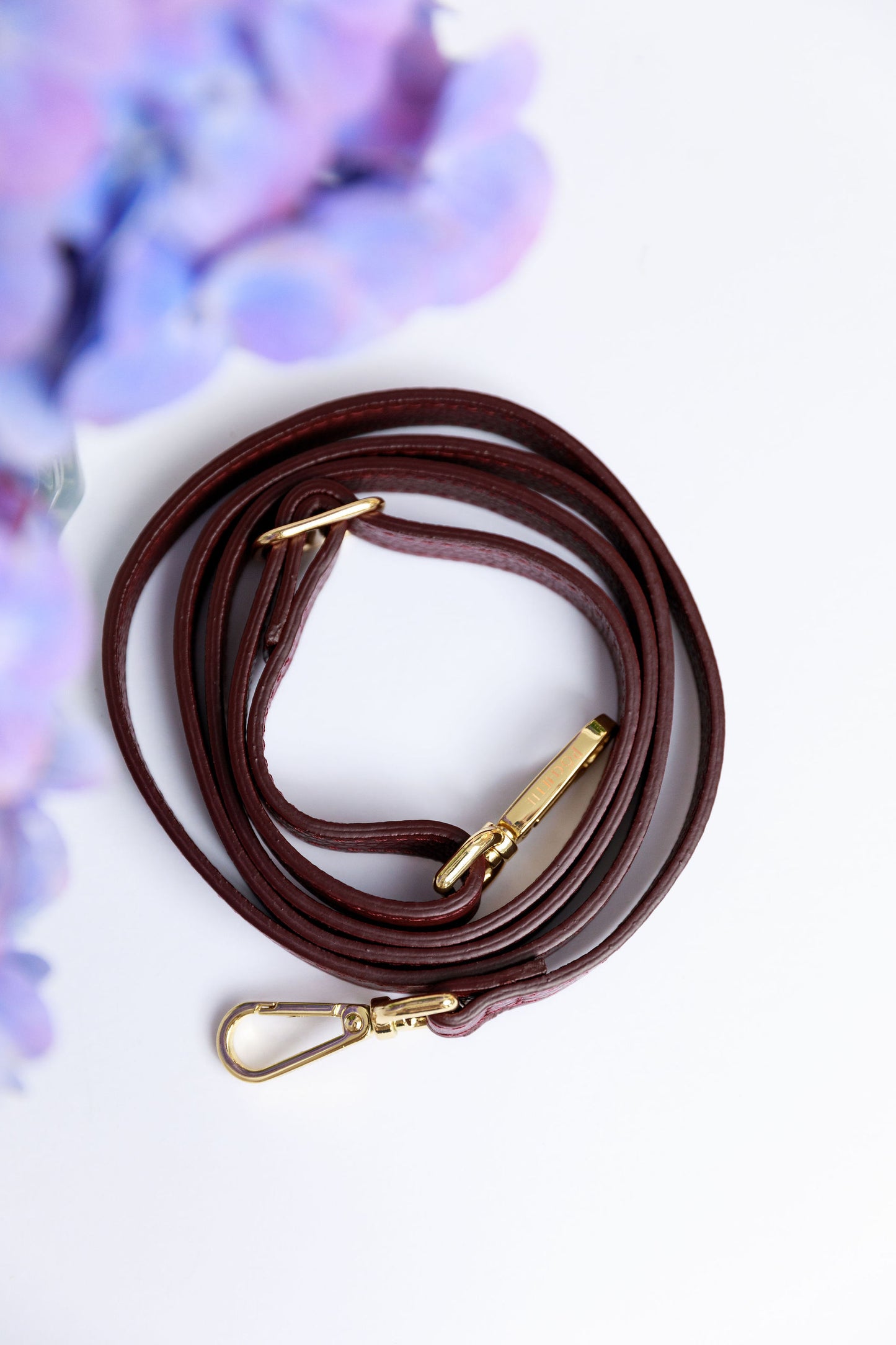 A burgundy thin leather strap on a white surface with a blurred hydrangea in the foreground.