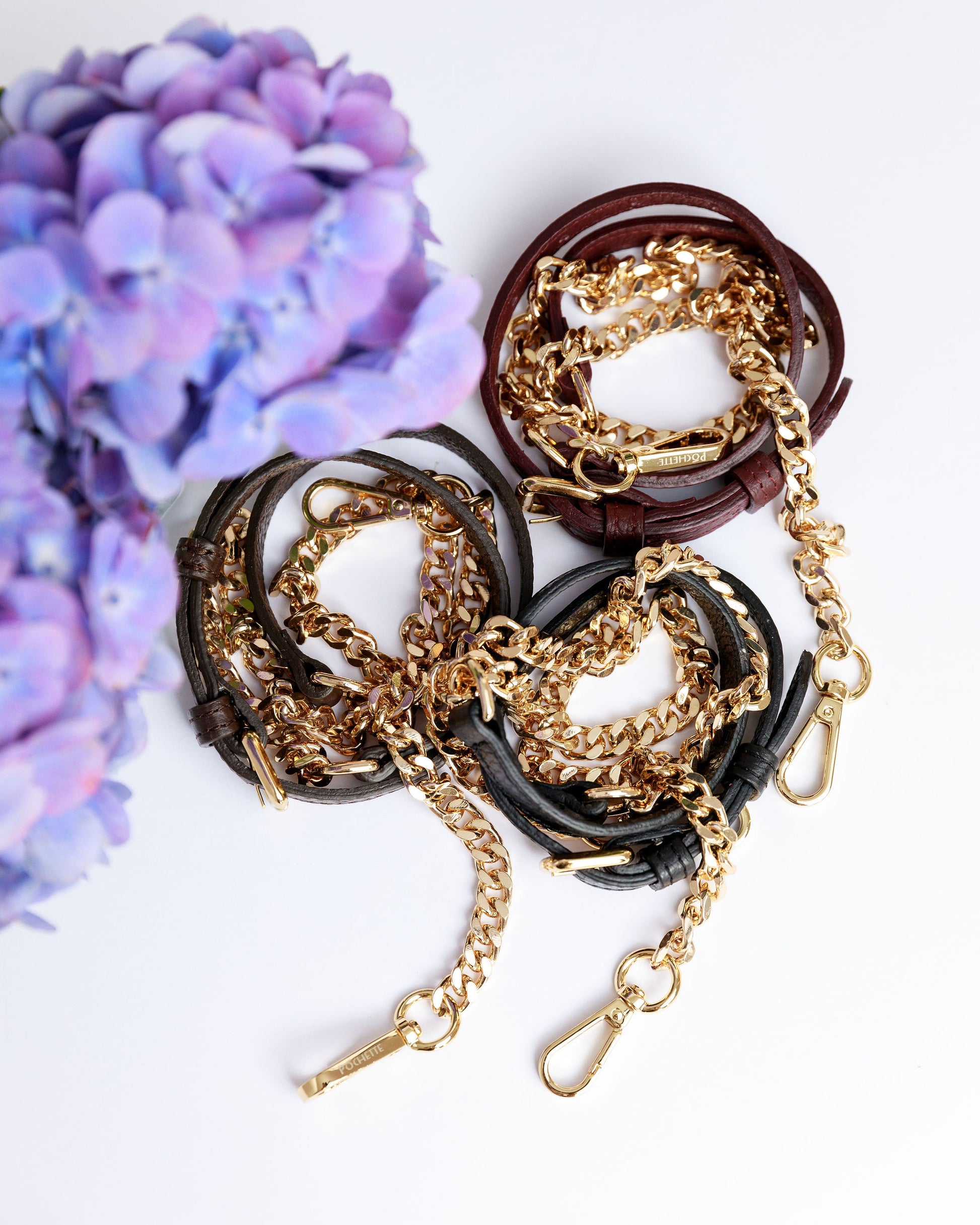 Black, chocolate brown and burgundy leather and chain straps on a white surface with a blurred hydrangea in the foreground.