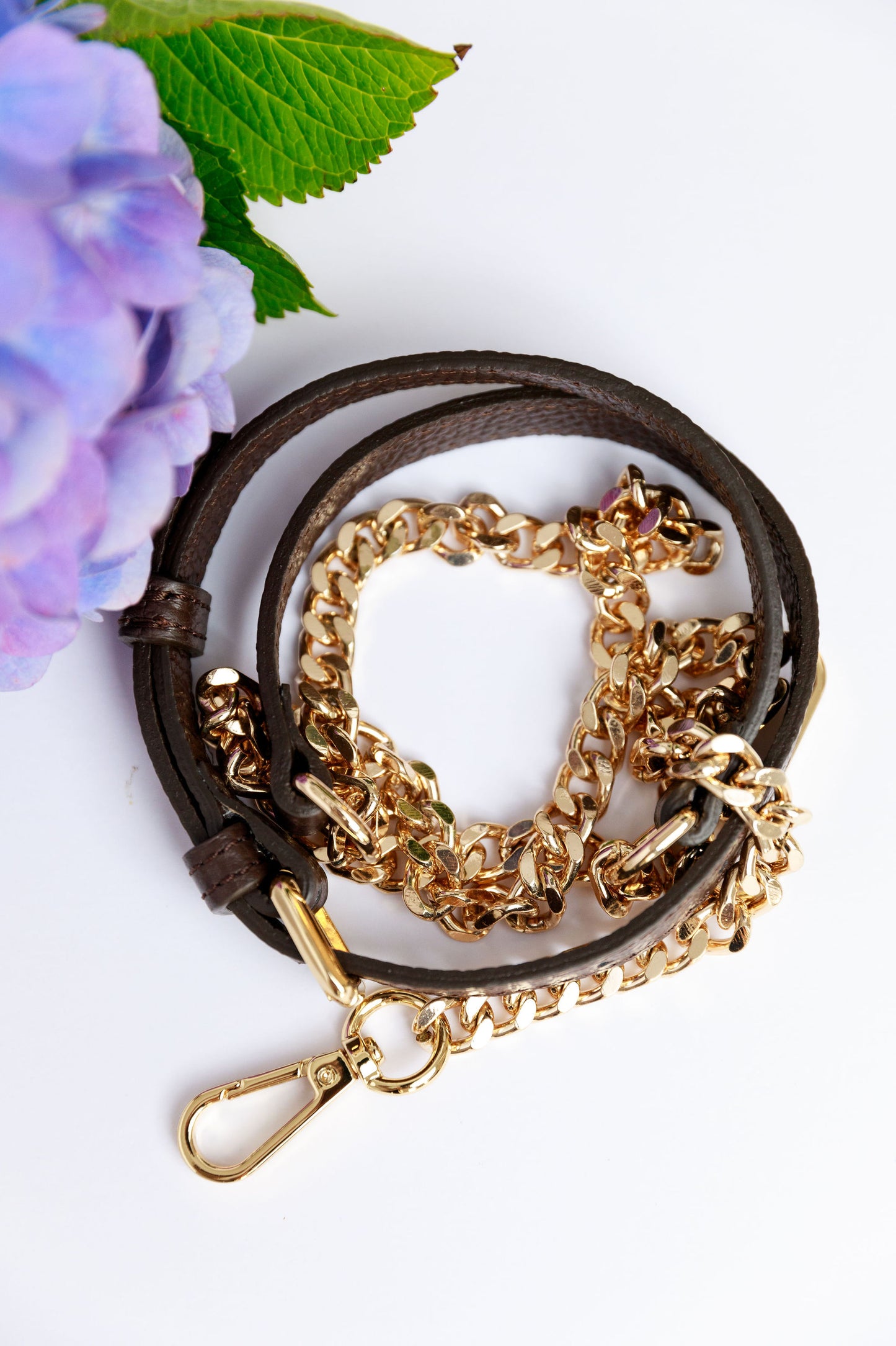 A chocolate leather and chain strap on a white surface with a blurred hydrangea in the foreground.